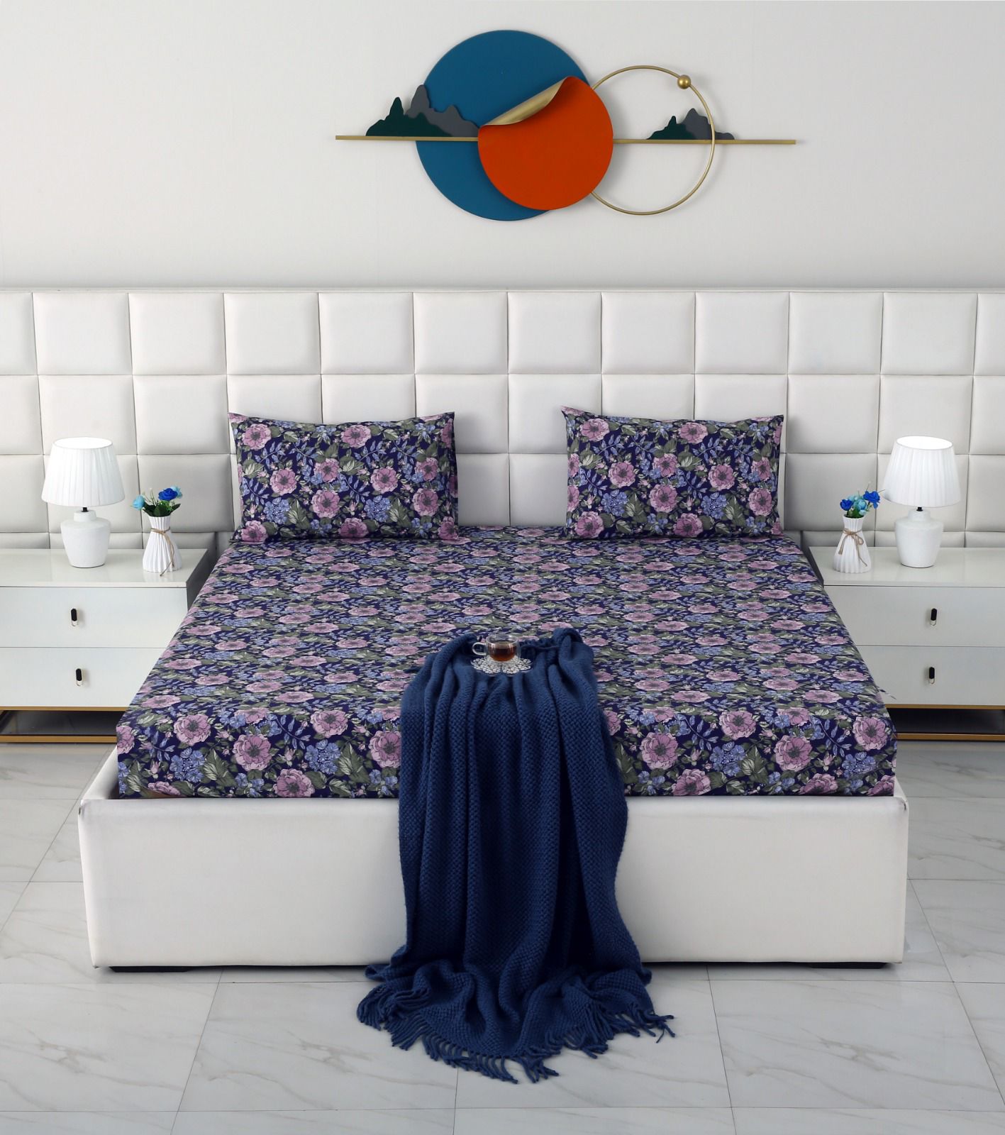 A neatly made bed with a floral-patterned bedding set in purple, pink, and blue, styled with a dark blue throw blanket. The bed is set against a white tufted headboard with matching white bedside tables and lamps, and a modern circular wall art piece hangs above.