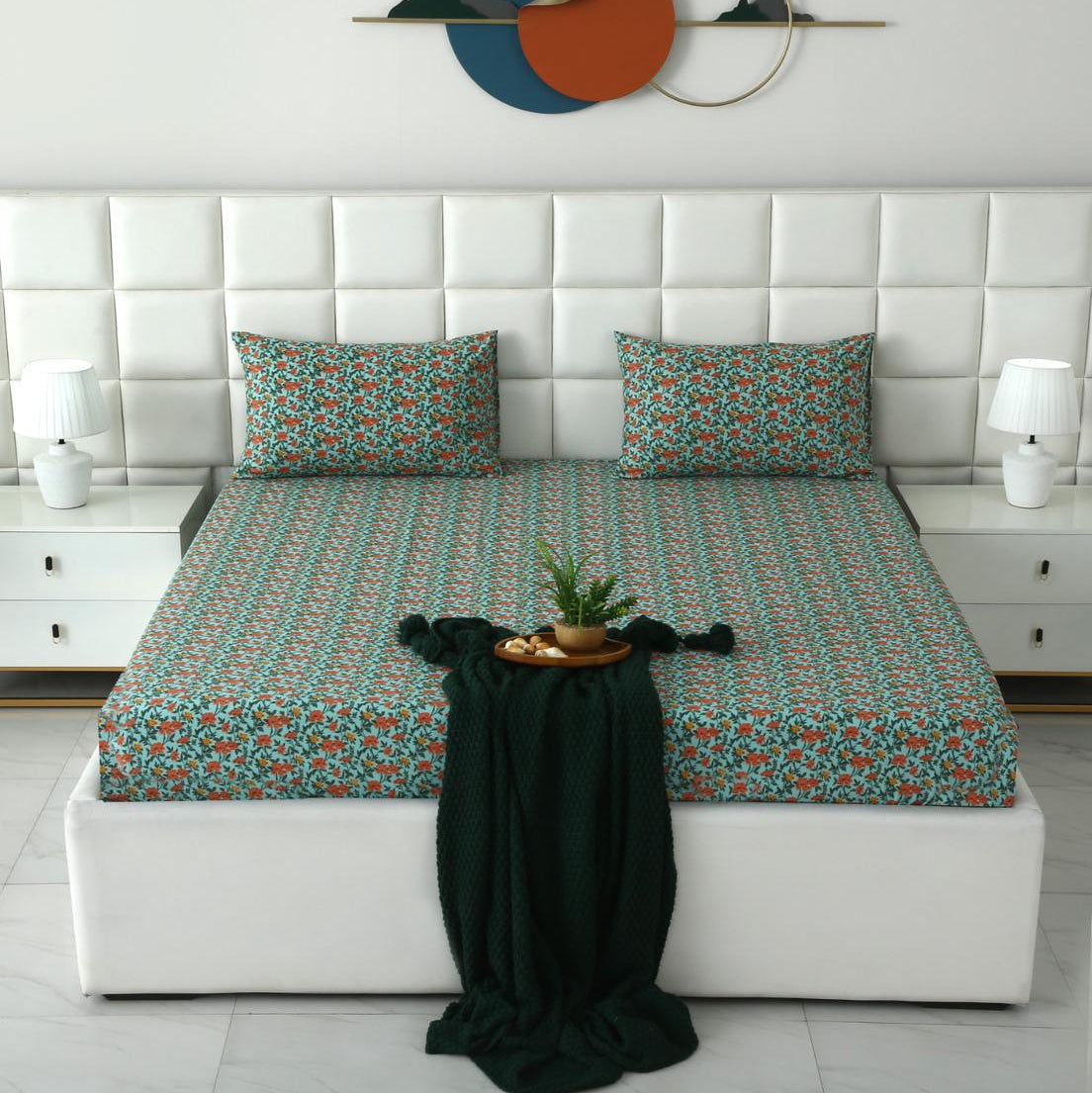 A neatly made bed with a teal and orange floral-patterned sheet and matching pillowcases, set in a modern bedroom with a white tufted headboard, bedside tables, lamps, and a dark green throw blanket.