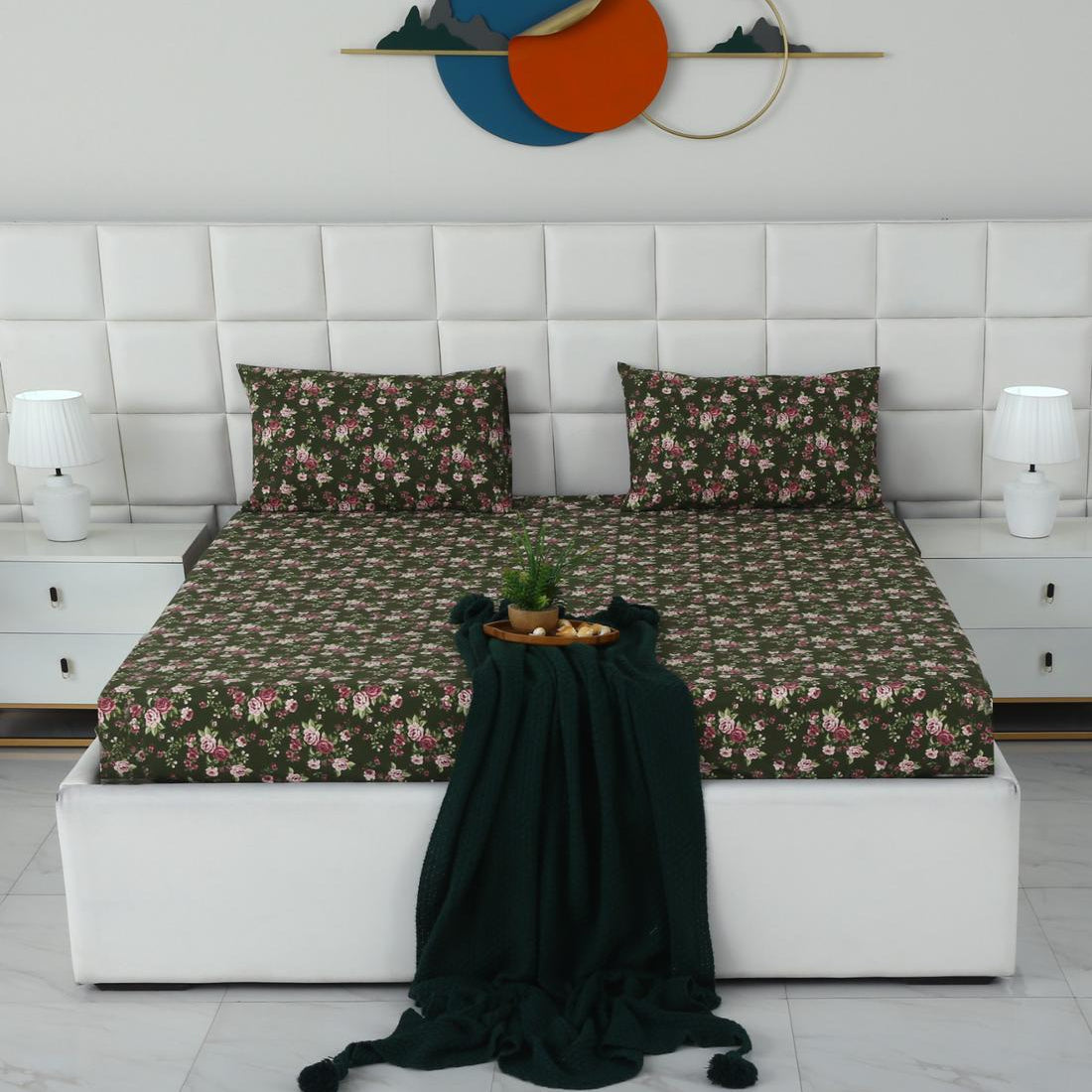 A neatly made bed with a dark green floral-patterned sheet and matching pillowcases, styled in a modern bedroom setting with a white tufted headboard, bedside tables, lamps, and a dark green throw blanket draped in front.