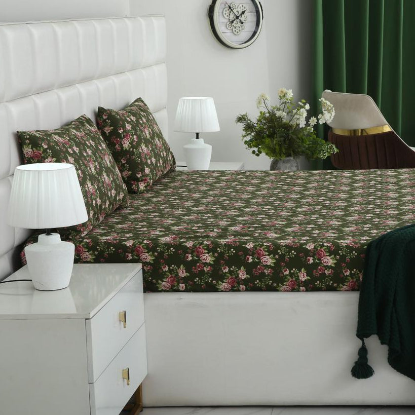 A bed dressed in a dark green floral-patterned sheet and matching pillowcases, styled in a modern bedroom with a white upholstered bed frame, bedside tables with white lamps, green curtains, and neutral-toned chairs.