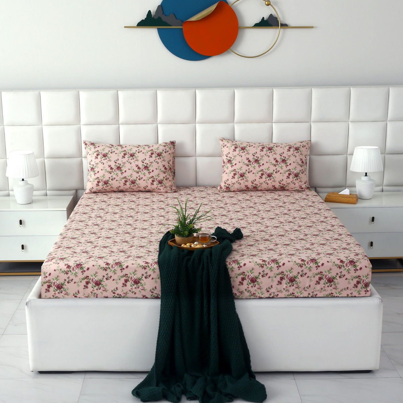 A neatly made bed with a pink floral sheet and matching pillowcases, set in a bright, modern bedroom with a white tufted headboard, white nightstands, and decorative wall art.