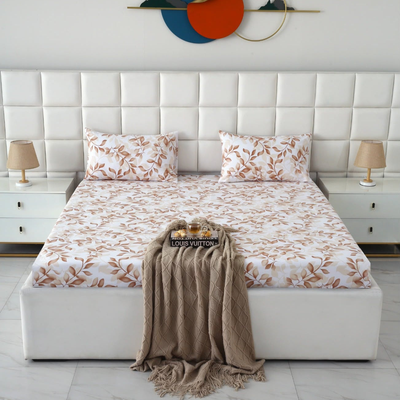 A neatly made bed in a modern bedroom setting, featuring a white tufted headboard, matching white bedding with a brown leaf pattern, two bedside tables with lamps, and a beige throw blanket draped over the foot of the bed.